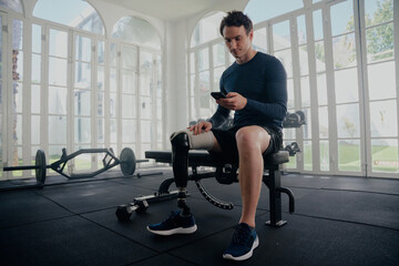 Paralympic athlete on his mobile in the gym training. Man with prosthesis sitting on bench looking at mobile phone .