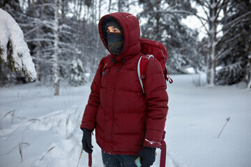 Picture of male hiker in red winter coat with hood wearing black snood scarf up to his nose and gloves, with backpack, standing in the middle of snowy forest, lost, holding smartphone in hands