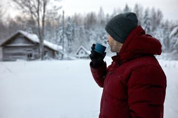 Fototapeta na wymiar Side view of male in red winter coat drinking tea from blue thermos cup standing against old wooden rural houses, snowy spruces, pines, looking, aside, traveling far away from big city