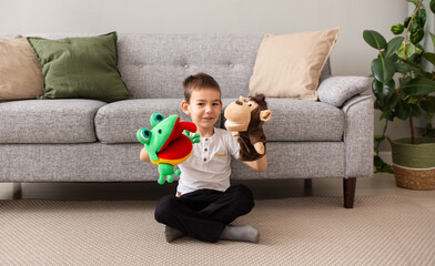 a Caucasian little boy is sitting on the carpet near the sofa and playing a puppet theater with a...