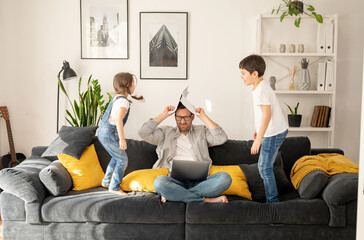 Annoyed father sitting with laptop with eyes closed, covering head with work papers while his two kids are jumping and shouting around. Irritated freelancer parent is babysitting and working