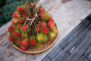A large bunch of red rambutan in a bamboo tray is placed on a wooden table. Looks delicious to eat rambutan, fresh fruit from the tree in Thailand. Leave space for text