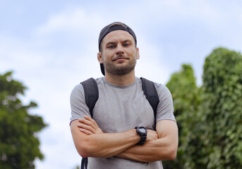 Adult man posing with crossed arms outdoors. Backpacker man in cap with a backpack.