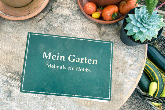 Auf einem verwitterten Holztisch liegt ein grünes Buch mit dem Titel "Mein Garten - Mehr als ein Hobby", drumherum liegt gemüse und pflanzen