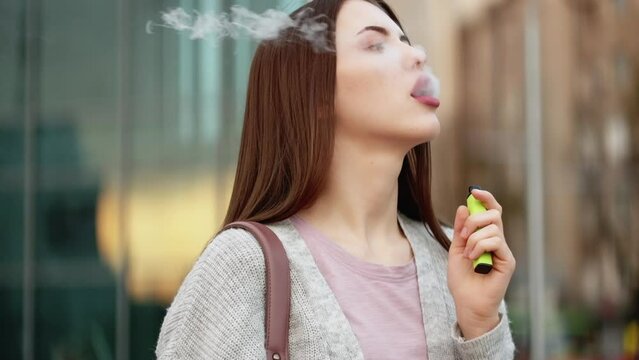 Vaping woman. Electronic cigarette. Nicotine addiction. Attractive elegant young female smoking outdoors exhaling steam at city street.