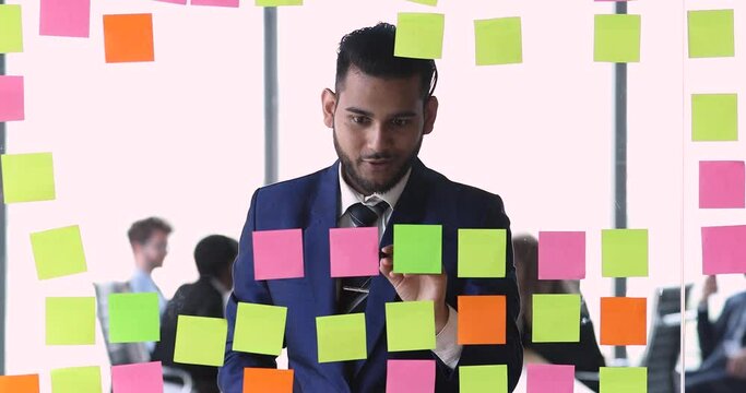 Smiling millennial businessman organize workflow process use partition of tasks by kanban methodology. Smart young man developer make planning visualization of project by sticking papers on glass wall