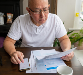 A senior man sitting by a table calculating the raising cost of energy and tax bills. Inflation and...