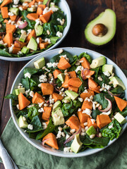 Avocado, roasted sweet potato, spinach, fried onions, pine nuts, feta cheese healthy salad in plate on wood background. Closeup, soft selective focus