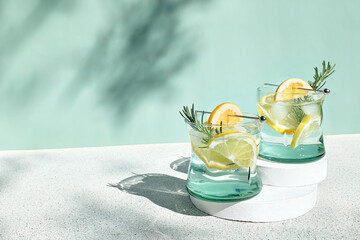Summer refreshing lemonade drink or alcoholic cocktail with ice, rosemary and lemon slices on pastel light green surface. Fresh healthy cold lemon beverage. Water with lemon. - obrazy, fototapety, plakaty