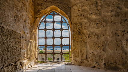 The window opening in the ancient Citadel of Qaitbay is barred. The texture of rough stone walls is visible. Shadows on the tiled floor. In the distance - blue sky, silhouettes of buildings in Egypt. 