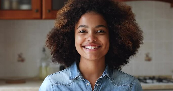 Close up head shot beautiful African teenage girl standing alone in home kitchen smile look at camera, having wide toothy charming smile, natural afro curly hairs poses indoors. Gen Z portrait concept