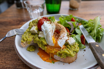 Sourdough toast, poached eggs, avocado pulp and fresh vegetables on plate in cafe, close up, breakfast time