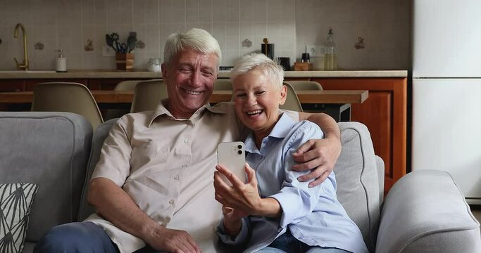 Older smiling happy grey haired couple sit on sofa at home, looking at cellphone screen watch funny on-line videos, elderly spouses have fun use modern smart phone. Leisure with wireless tech concept