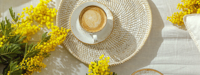 Branches of yellow mimosa flowers on white linen bed sheets, cup or coffee. Greeting for International Womens Day on March 8th, mother’s day or spring concept.