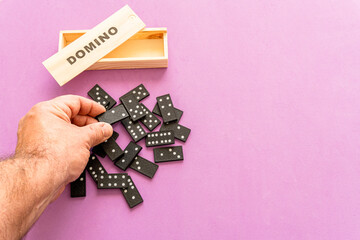 Male hand on the tiles of a game of dominoes disordered on a pink background.