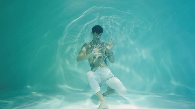Underwater shot of young man entangled in chains. Man screams and tries to get out of blue water column with glare. Emotions of suffering, captivity of chains. Subaquatic shot, close up in slow motion