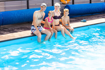 Caucasian family having fun by their swimming pool. Happy young family splashing water with hands and legs while sitting on edge of swimming pool. Kids with parents playing outdoors