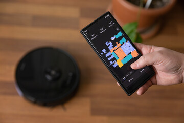 Close up of man holding mobile phone with remote control for robot vacuum cleaner, searching room...