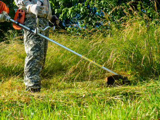 A man dressed in work clothes using a gasoline mower cuts long grass. Summer. rural area, maintenance of the territory