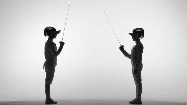 Silhouettes of two female swordsmen fighting a duel. Athletes cross rapiers and deliver attacking blows. The girls pose in a dark studio on a white background in a spotlight spot. Slow motion.