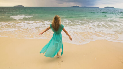Beautiful long-haired woman walks to the stormy cloudy ocean on sand beach. Girl in blue swimsuit dress tunic. Concept rest in sea, tropical resort coastline traveling tourism summer beach holidays
