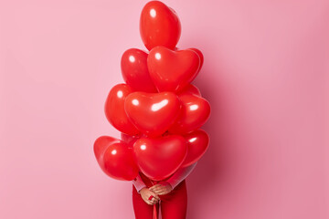 Unknown woman hids behind big bunch of heart shaped balloons celebrates Valentines Day poses against pink background. Unrecognizable female model prepares for celebration going to have party