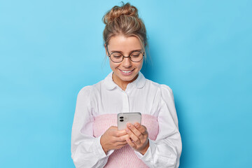 Pleased elegant woman wears formal white shirt round spectacles concentrated in smartphone checks received notification enjoys social networking via cellphone application isolated over blue background