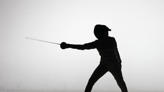 Silhouette of young woman swordsman throwing attacking blows with sword. Sports training in fencing school. Girl athlete posing in dark studio on a white background in spot of spotlight. Slow motion.