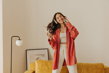 Cheerful caucasian woman is listening to music and dancing on sofa in bright cozy living room. Photo of entertaining dark-haired beauty with headphones and phone in hands. Fashion, technology concept.