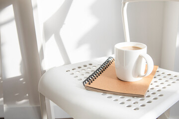 White cup with coffee, notebook on a white chair under natural light.