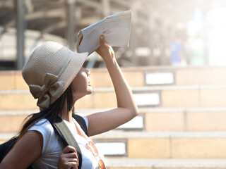Close up, side pose of Asian female tourist wearing hat and backpack using city map blocking sunlight on sunny hot day.
