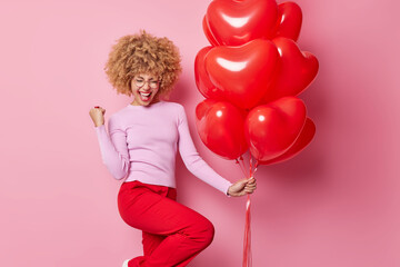 Upbeat curly haired woman clenches fist with joy happy to celebrate Valentines Day holds heart shaped balloons wears casual jumper and trousers isolated over pink background. Holidays and celebration
