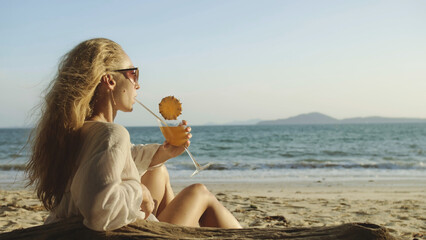 Fototapeta na wymiar Attractive woman sitting in white dress sunglasses, drinking pineapple cocktail Pina Colada. Beautiful beach, sea waves at sunset. Concept rest tropical resort traveling tourism happy summer holidays