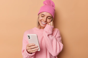 Gentle woman looks at smartphone happy to get message with pleasant content from lover smiles with teeth dressed in pink hat and jumper poses against beige background. Emotions and technology