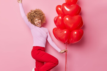 Happy celebration concept. Glad joyful woman dancs carefree with bunch of red heart balloons has upbeat mood during Valentines Day wears casual jumper and trousers isolated over pink background