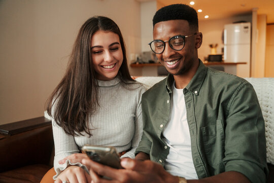 African American male sitting on sofa with caucasian female girlfriend holding cellular device texting