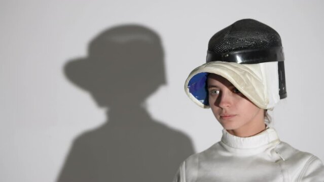 Portrait of a young woman fencer saluting with rapier, putting on helmet and getting into fighting position. An athlete poses in dark studio on white background with shadow. Slow motion. Close up.