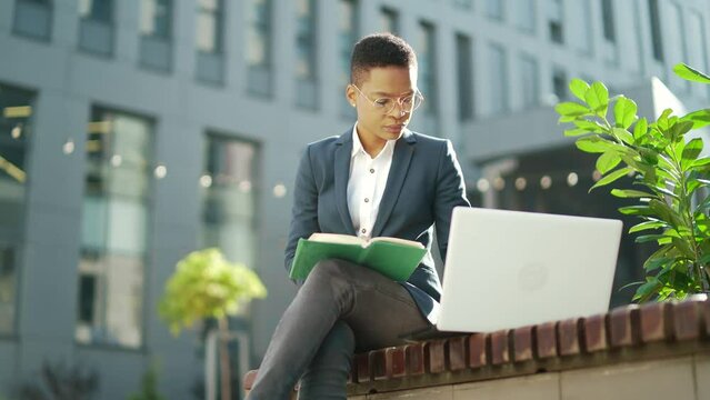 Young african american business female student sitting in a city park on bench with laptop and notebook studying online outdoors Woman freelancer in glasses learn working remotely in street e-learning