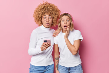 Shocked impressed young women use modern technologies stare wondered at camera hold smartphone listen radio online find out shocking news dressed in casual clothes isolated over pink background