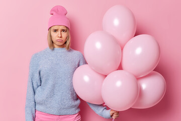 Fustrated unhappy blonde young woman looks sadly at camera holds big bunch of inflated balloons feels lonely during birthday celebration dressed in casual clothes isolated over pink background.