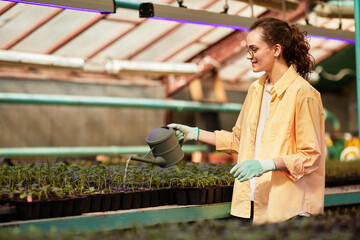 Young happy woman in gloves, shirt and eyeglasses watering sprouts while holding watering-pot over them during work in hothouse