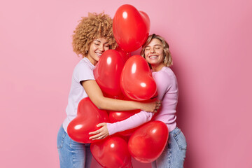 Two positive young women embrace bunch of inflated red heart shaped balloons prepare for Valentines Day enjoy romantic holiday dressed casually isolated over pink background. Romantic holiday