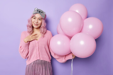 Obraz na płótnie Canvas Good looking pink haired girl feels thankful receieves heartwarming words of congratulation on her birthday wears sleepmask jumper and skirt poses with bunch of balloons isolated on purple wall