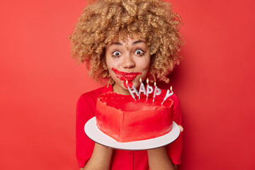 Surprised curly haired woman make mess eating big fancy heart shaped cake has mouth smeared with...