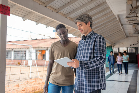 Hight school african student talking with his teacher in the hallway, other students talking in the background.