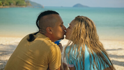 The love couple is holding a glass of blue curacao cocktail, on sea. Man with plait and blonde woman drink alcohol on sand beach in shadow by azure ocean of a tropical island. Summer holiday concept.