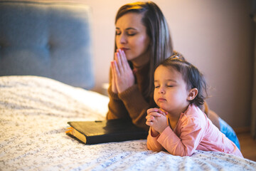 Mother and her daughter reading from bible and praying in their knees near the bed - 486945075