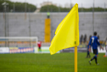 Yellow flag at one corner of football stadium and soccer corner of a soccer field with match action in the background.