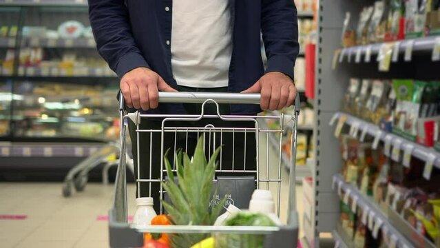 Young male consumer holding trolley and walking in supermarket while shopping spbd. Close-up view of Caucasian man moves cart filled with organic fruits, vegetables and walks along row, looks at goods