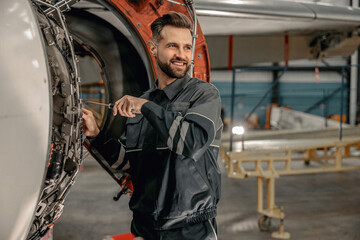 Joyful bearded man maintenance technician using screwdriver and smiling while repairing airplane at...